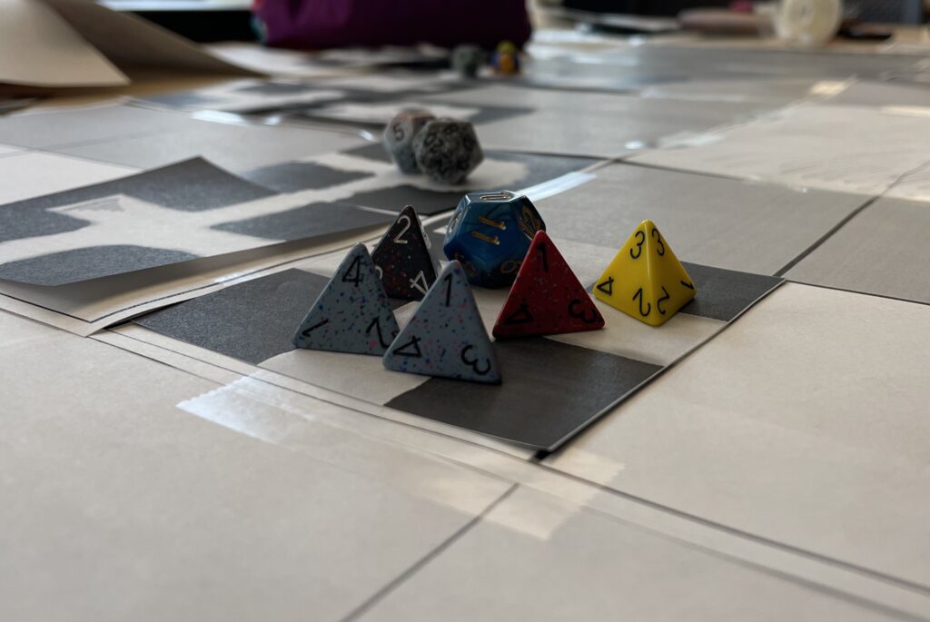 Six dice sit on a black & white map composed on square paper pieces. Five of the dice are four-sided, while one is twelve-sided. In the background, more map tiles and dice are visible.