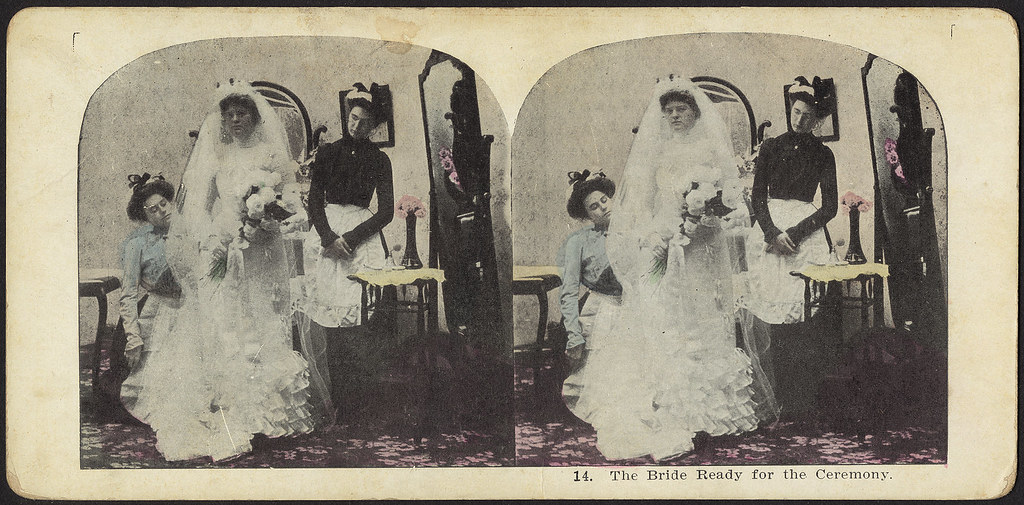 A stereograph from around 1900 showing a bride getting ready. A maid and seamstress help the bride dress while she poses, expressionless, holding a bouquet of flowers.
